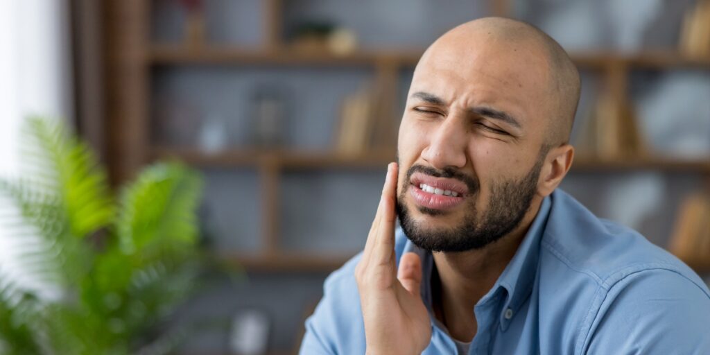 Young man in a blue shirt clutching his cheek in a living room, grimacing from severe tooth pain and seeking urgent dental care with a pained, distressed expression